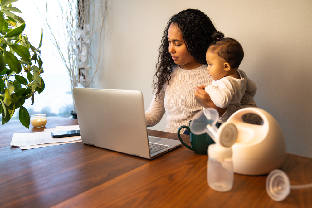 A mother holds her infant while working at a laptop computer with her breast pump nearby