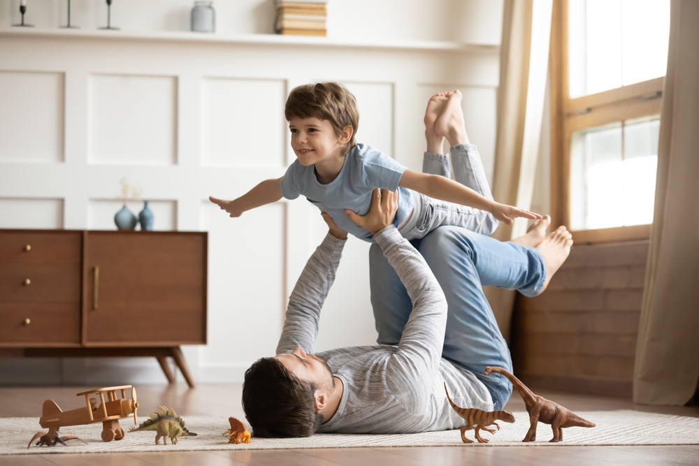 A father lies on his back on the floor and lifts his young son into the air